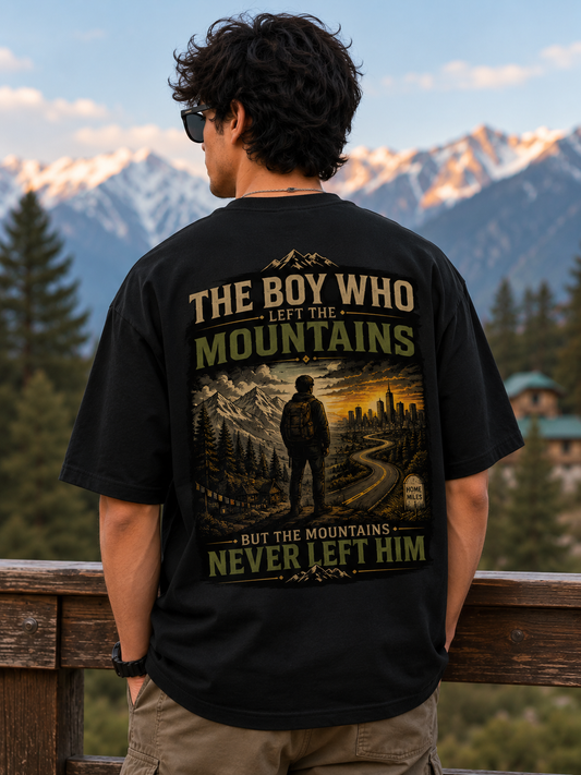 Person wearing a black t-shirt with a mountain-themed design, standing in front of a scenic mountain landscape.